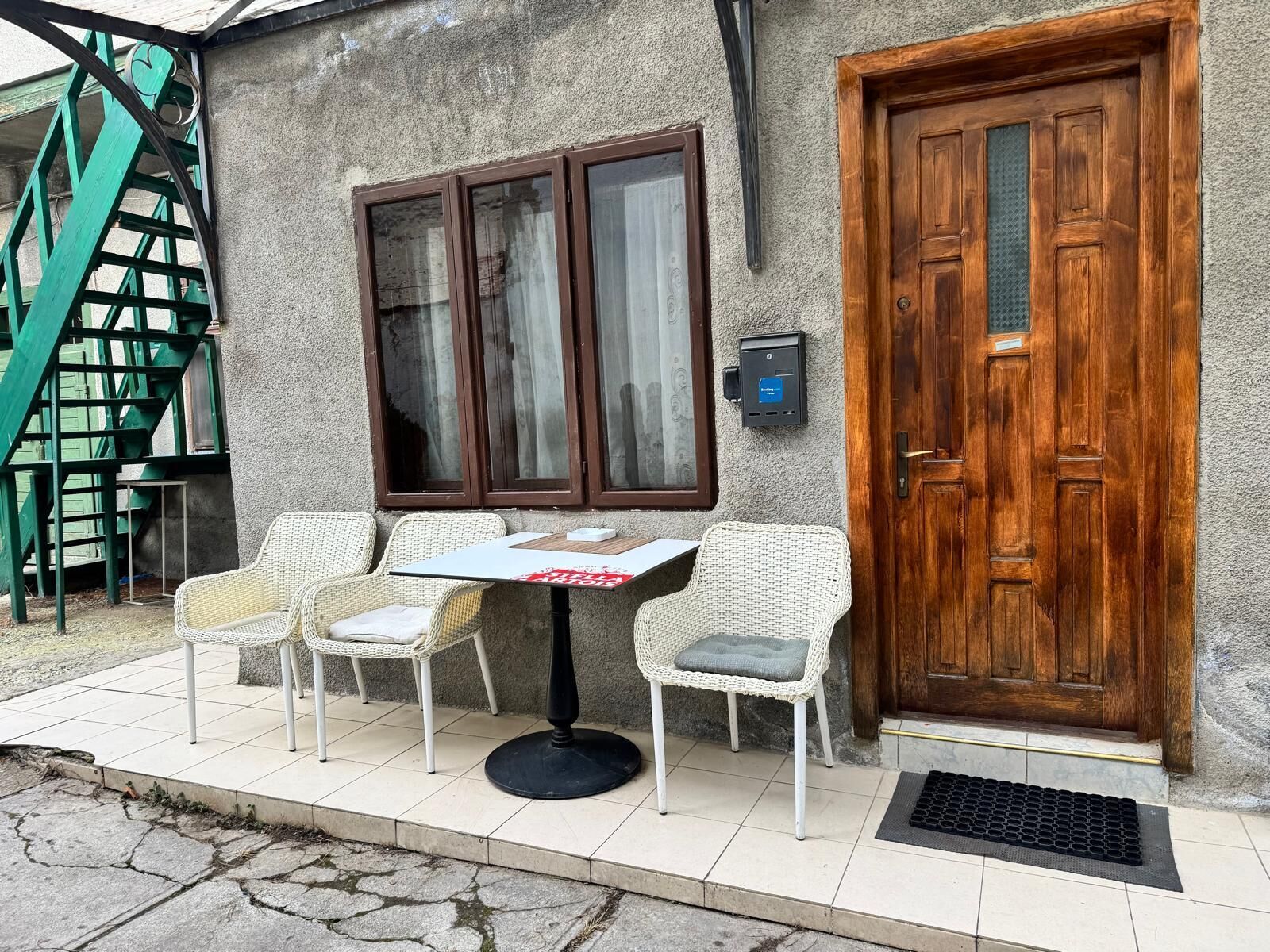 Um quarto com porta de madeira, janela com cortinas, uma mesa com cadeiras e um lance de escadas verdes.