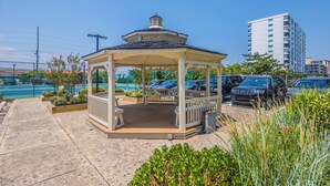 Un kiosque doté d’un toit en tuiles et d’un banc, entouré de verdure et bordé d’un parking où sont garées des voitures.
