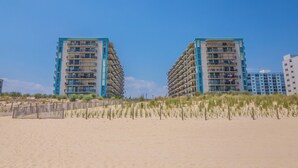 Deux immeubles de grande hauteur situés en bord de mer, dotés de balcons, une plage de sable au premier plan et un ciel bleu dégagé.