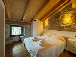 A bedroom with a stone wall, wooden beams, a window, a bed, and bedside tables.