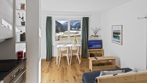Une pièce comprenant un lit, un téléviseur, un bureau avec des chaises, une fenêtre avec des rideaux et une étagère avec des plantes.