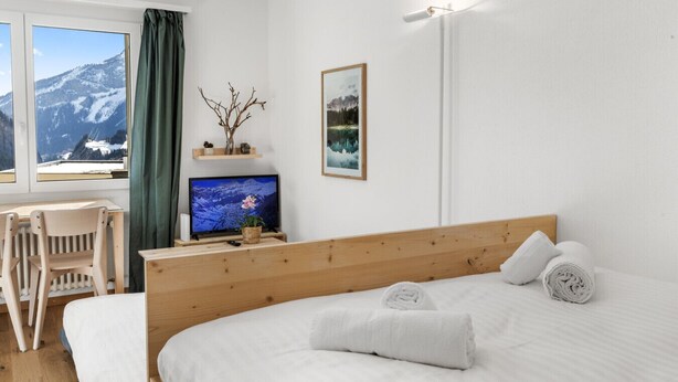 Une chambre avec une tête de lit en bois, un lit recouvert de draps blancs, un téléviseur posé sur un meuble en bois, une chaise, une fenêtre donnant sur les montagnes et un luminaire fixé au mur.