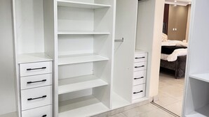 A walk-in closet with open shelves and drawers, a bed with a headboard visible in the background, and a hanging rack.