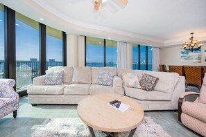Un salon moderne avec un grand canapé d’angle beige, une table basse ronde et une vue sur la ville grâce à de grandes fenêtres.