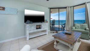Una sala de estar con televisor, una mesa de centro de madera, un sofá y vista al océano a través de un ventanal corredero.