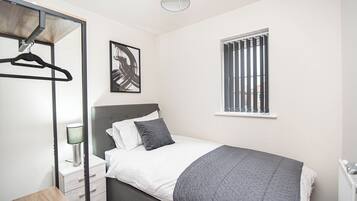 Une chambre à coucher comprenant un lit, une table de chevet, une lampe suspendue, une fenêtre avec des stores et une œuvre d’art accrochée au mur.