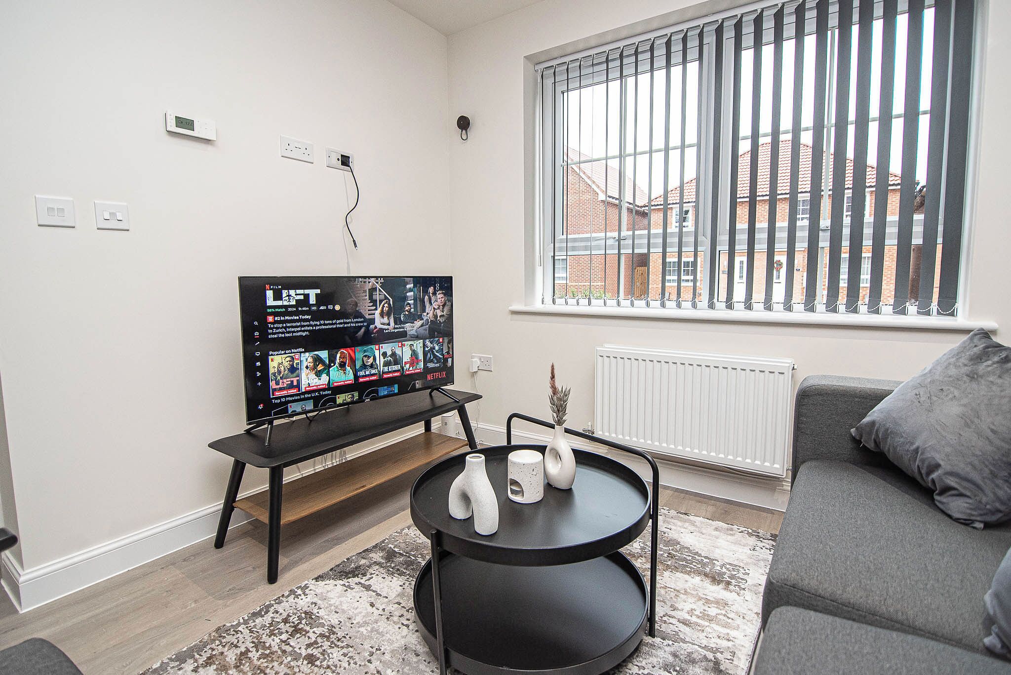 Een moderne woonkamer met een flatscreen televisie, een zwarte salontafel met vazen, een grijze bank en een raam met zonwering.
