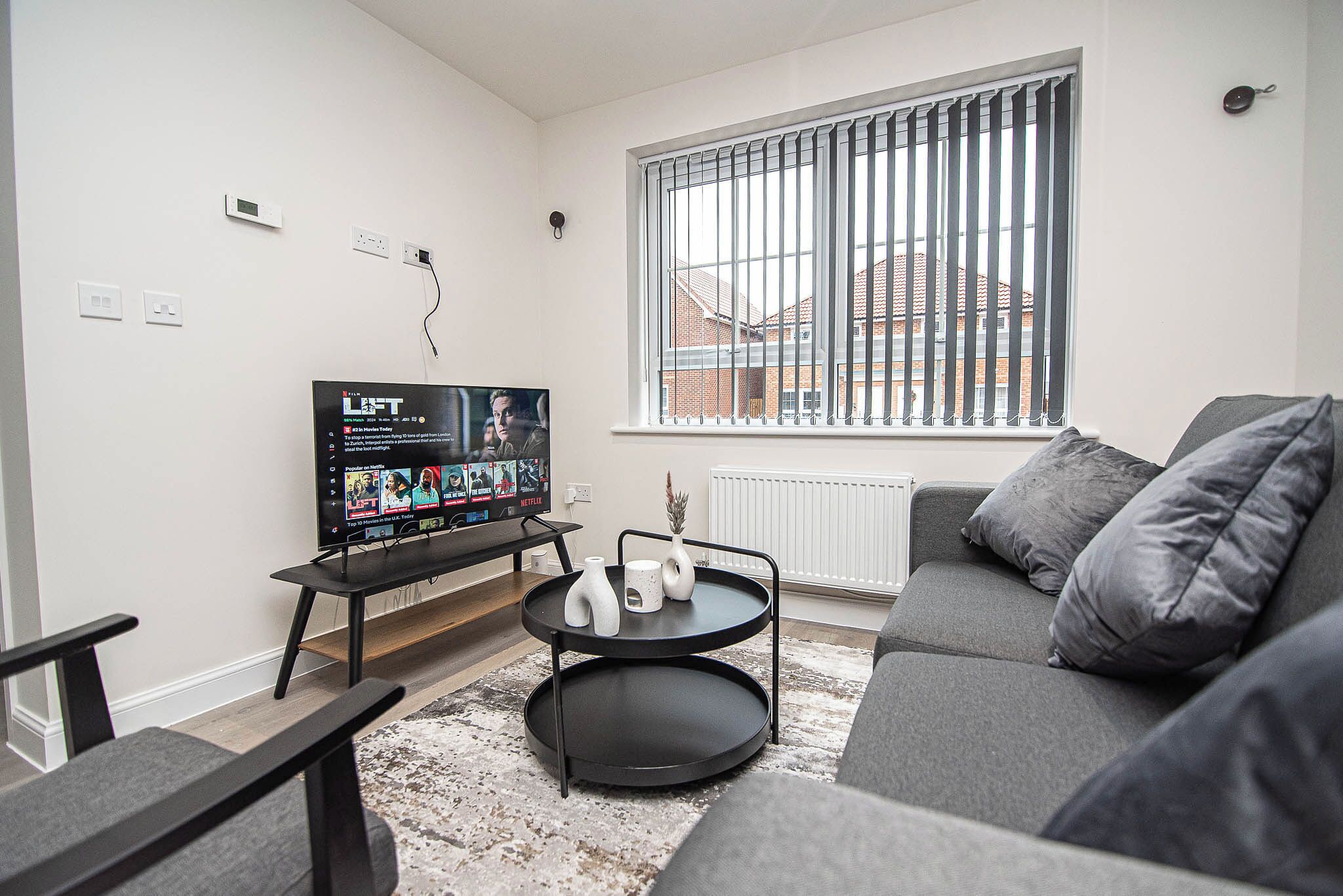 Ein modernes Wohnzimmer mit einem grauen Sofa, einem schwarzen Couchtisch mit Vasen, einem Flachbildfernseher und einem Fenster mit Jalousien.