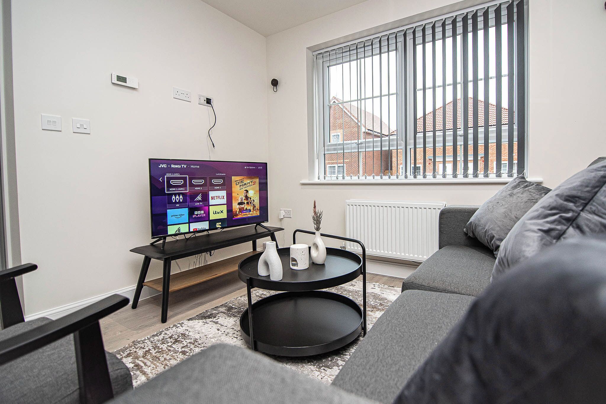 A modern living room with a flat-screen TV, a two-tiered black coffee table, a grey sofa, and a window with vertical blinds.