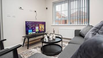 A modern living room with a flat-screen TV, a two-tiered black coffee table, a grey sofa, and a window with vertical blinds.