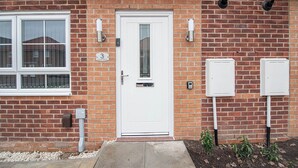 Une maison en briques avec une porte d’entrée blanche, une boîte aux lettres et deux boîtiers électriques sur le côté.