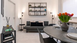 Een moderne woonkamer met een grijze bank, een ronde eettafel, een boekenkast en een vaas met bloemen.
