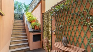 En smal udendørs korridor med en trætrap, et bord med en mønstret vase og en grøn espalier-væg.