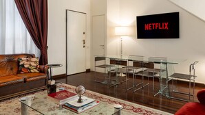 En moderne stue med en lædersofa, et glasbord med bøger og en globus, et spisebord i glas med metalstole og et vægmonteret tv med teksten 'NETFLIX'.