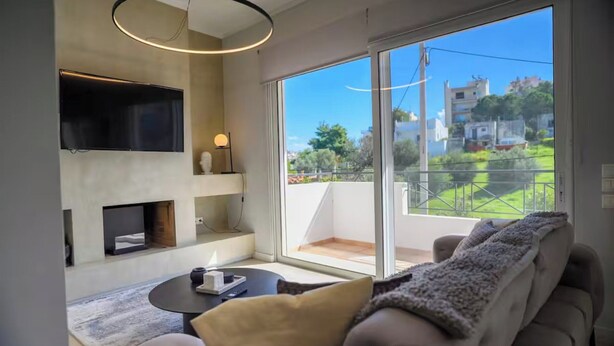 A modern living room with a flat-screen TV, a fireplace, a sofa, and a large sliding glass door to a balcony. - Green Hill Recreational Luxury (Iraklio)
