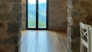 Une pièce rustique aux murs de pierre, dotée d’un plancher en bois, d’une grande fenêtre offrant une vue sur des collines verdoyantes et d’une chaise blanche.