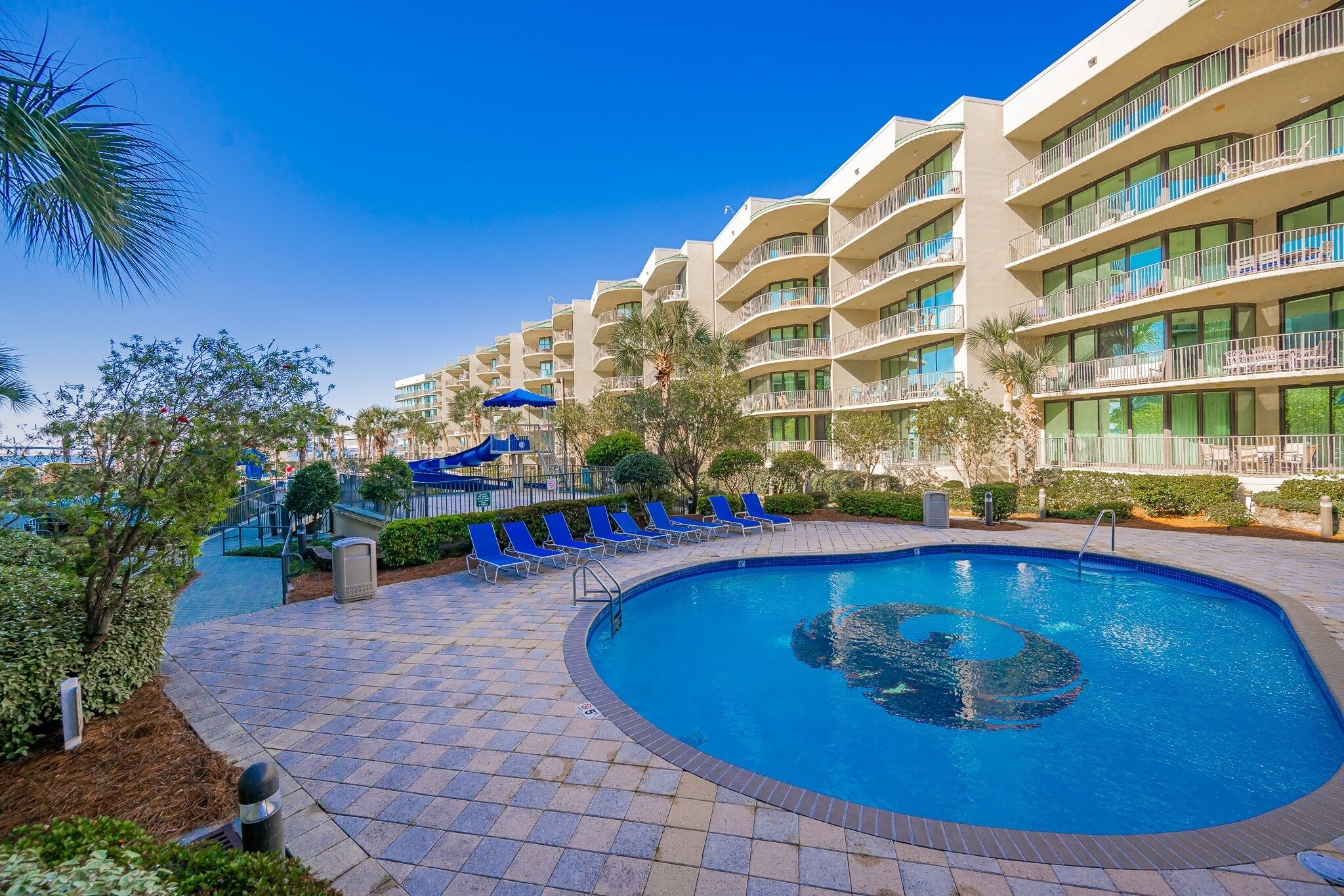 A hotel with a swimming pool, lounge chairs, and a clear blue sky.