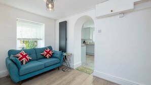 Un salon avec un canapé de couleur turquoise, deux coussins ornés du motif Union Jack, une petite table d’appoint et une fenêtre avec des stores.