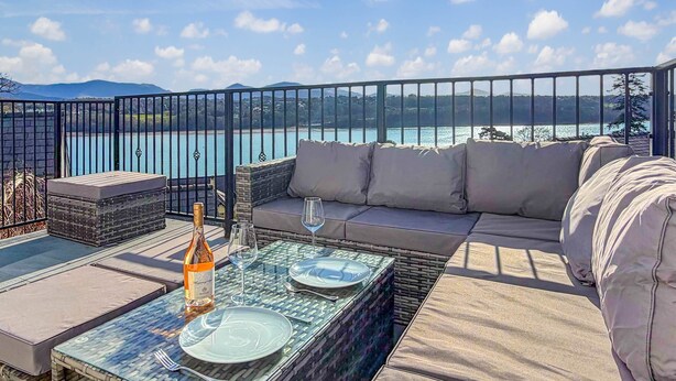 Une terrasse dotée d’un canapé, d’une table à manger et de chaises, offrant une vue sur un plan d’eau et des montagnes.