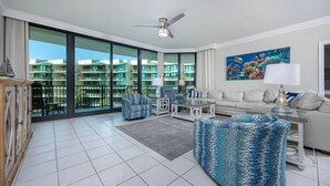 A spacious living area with a large window, a ceiling fan, a sofa, a coffee table, and a patterned armchair. - Phoenix On The Bay 1328 3 Bedroom Condo (Orange Beach)