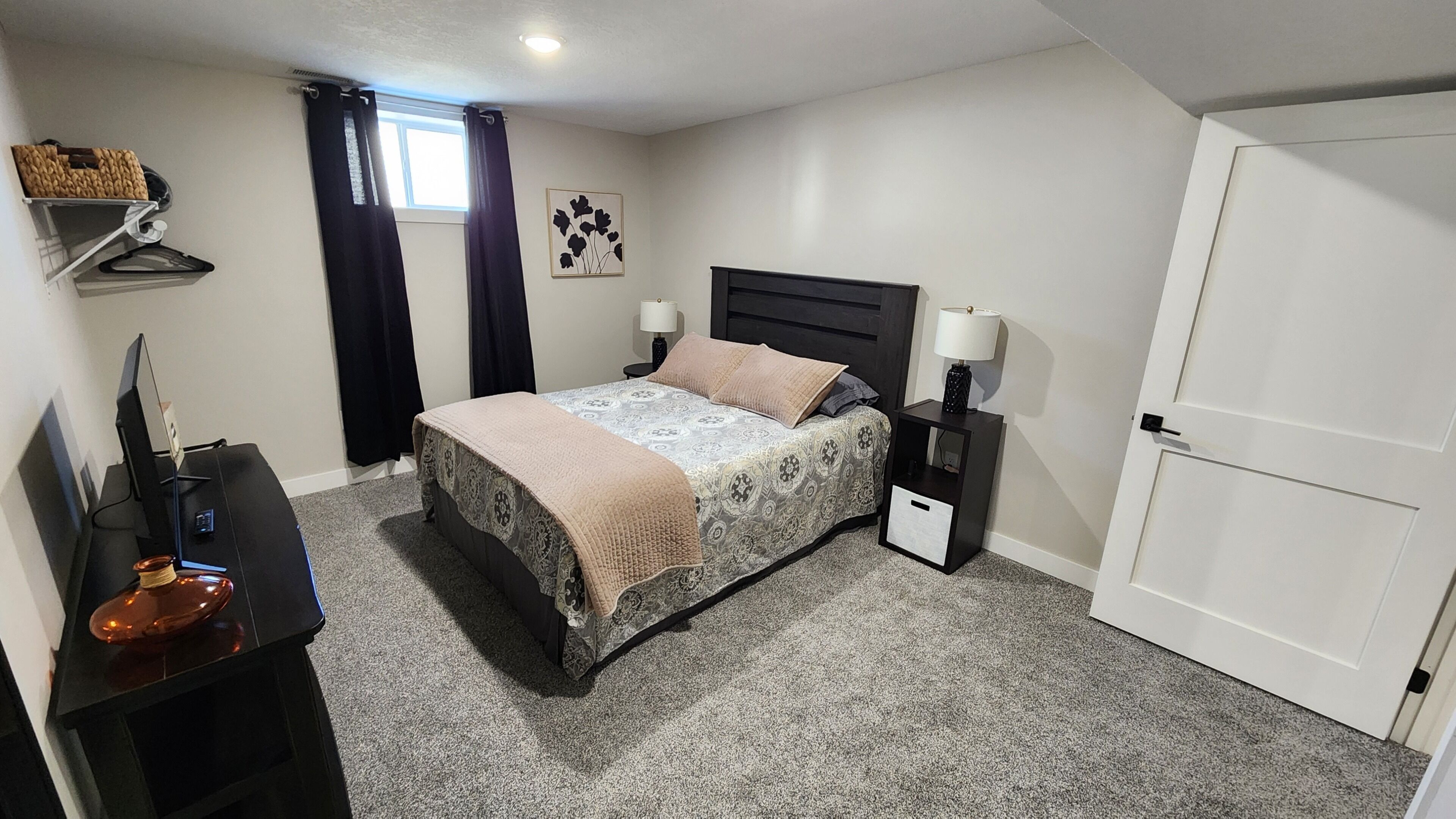 A bedroom with a bed, bedside tables, a desk, a chair, a lamp, a window with curtains, and a door.