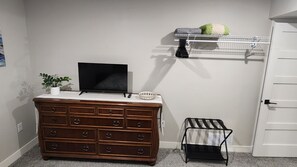 Quarto com cômoda de madeira, televisão em cima, um pequeno banquinho, prateleira com planta e uma porta.