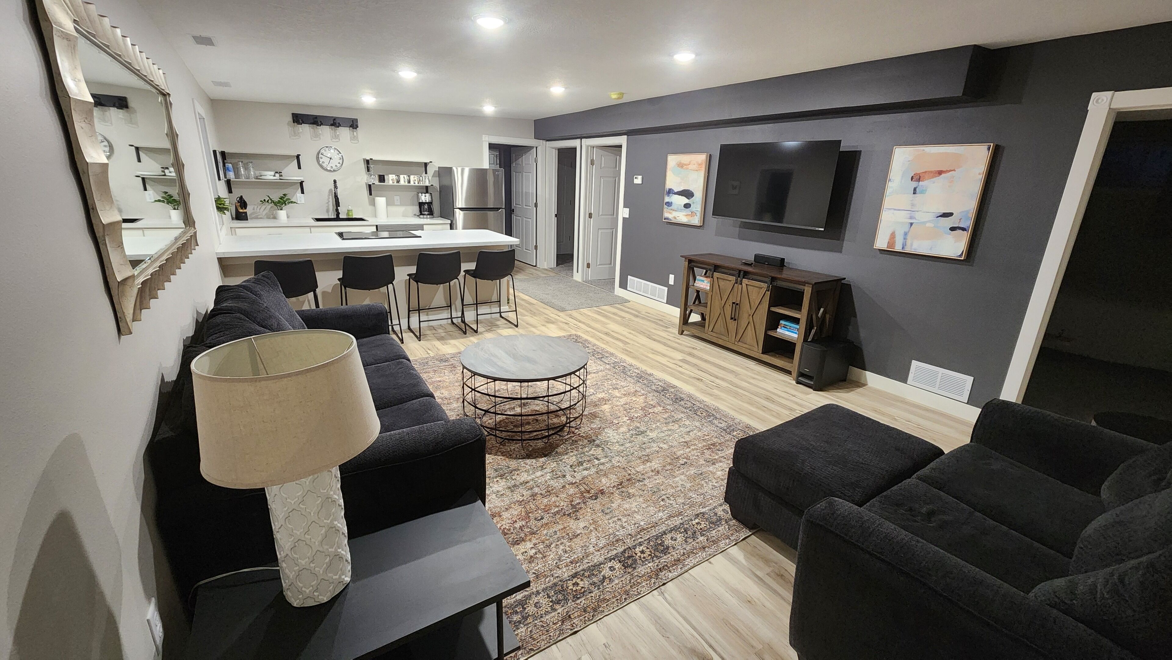 A modern living room with a sofa, ottoman, and a round coffee table. There is a kitchen area in the background with a refrigerator and a dining table.