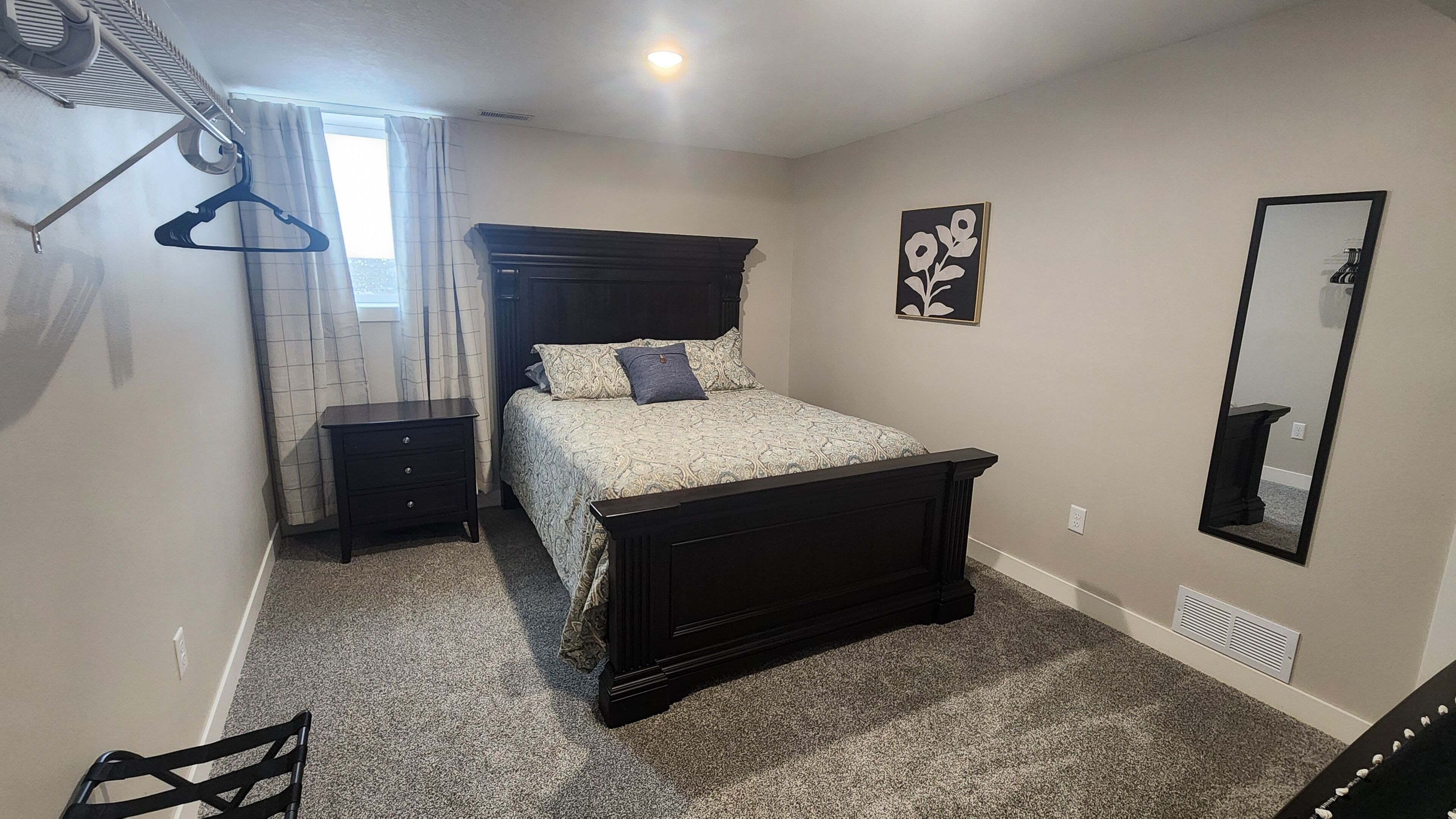 A bedroom with a bed, a nightstand, a dresser, a hanging coat rack, a wall-mounted mirror, and a window with curtains.