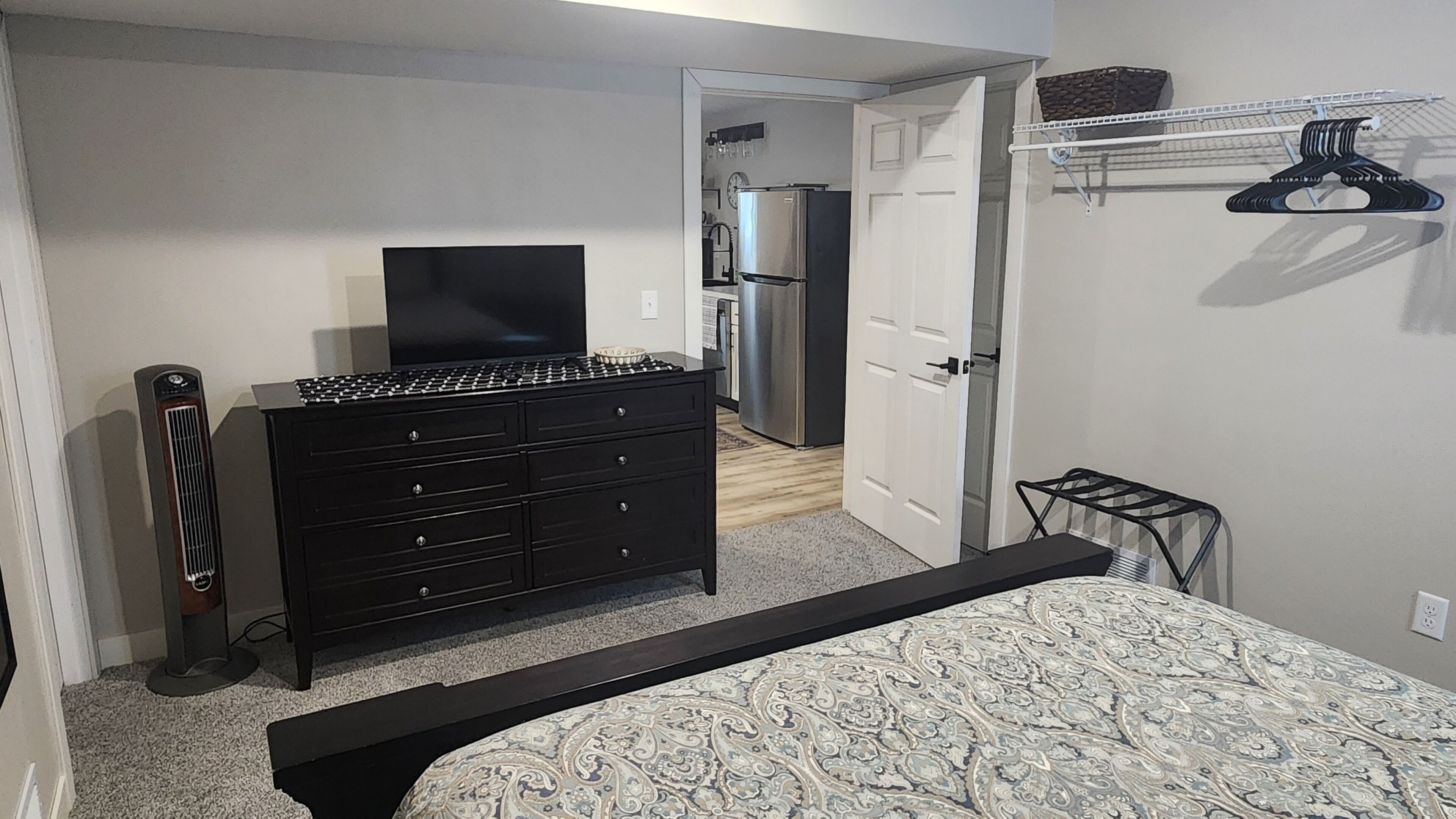 Une chambre à coucher comprenant un lit, une commode, une télévision et une porte donnant sur la cuisine.