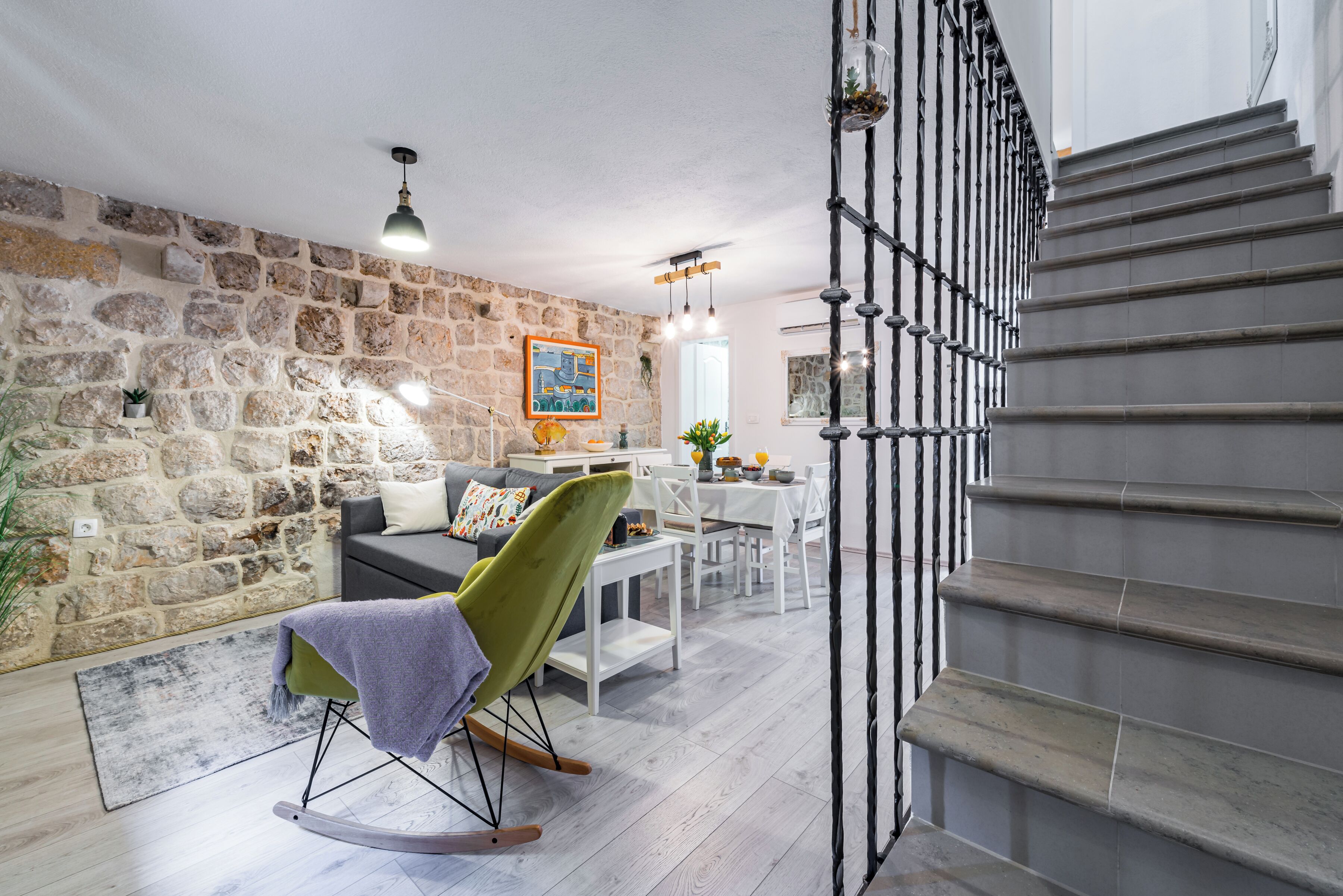 A modern living room with a stone wall, a green rocking chair, a dining area, and a staircase.