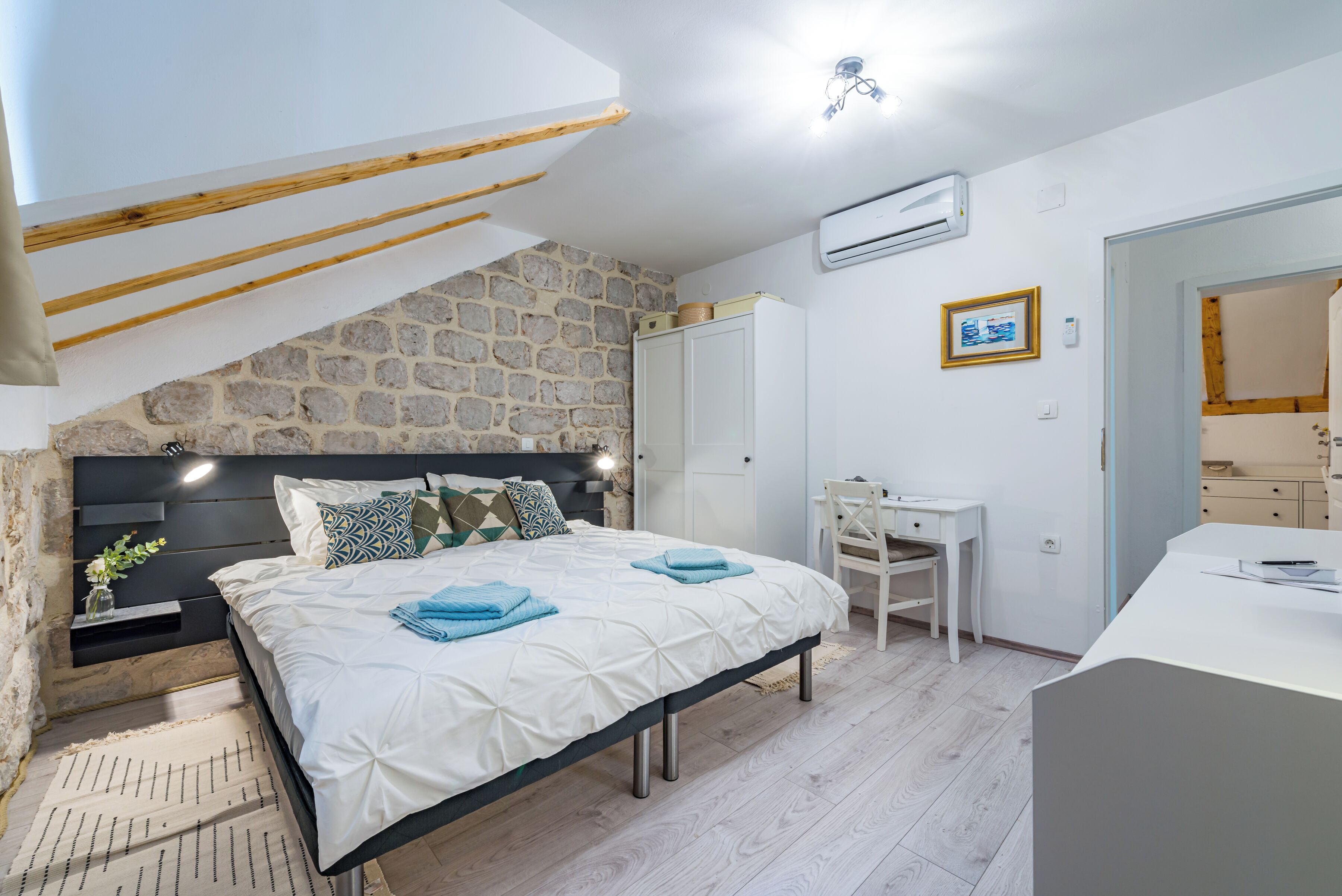A bedroom with a stone wall, a bed with white bedding, a desk, and a chair.