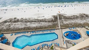 A swimming pool with blue water, surrounded by lounge chairs and umbrellas, with a beach and ocean in the background. - Phoenix V 1209 2 Bedroom Condo (Orange Beach)