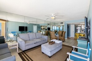 A living room with a sofa, ottoman, and chairs, a kitchen area with a refrigerator and counter, and a dining area with yellow stools. - Phoenix V 306 2 Bedroom Condo (Orange Beach)