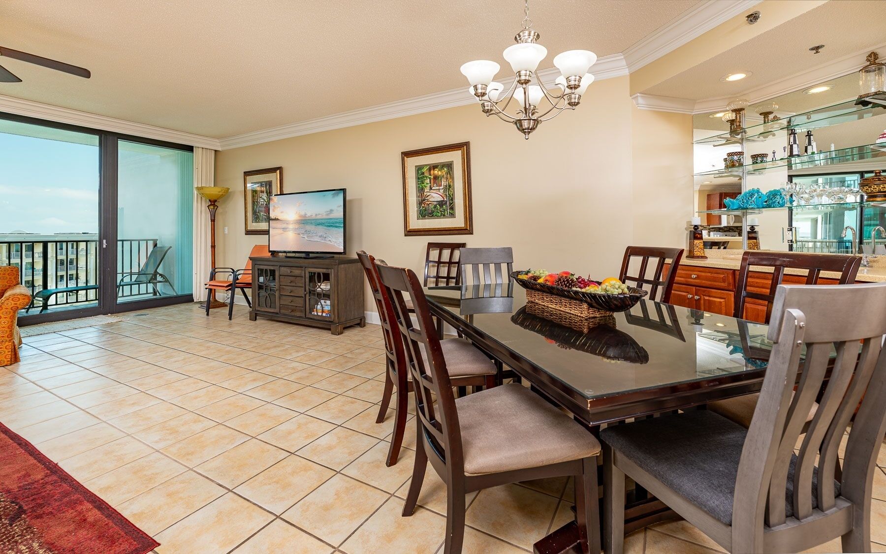 A spacious dining area with a glass-top table, surrounded by chairs, a television, and a balcony with outdoor seating.