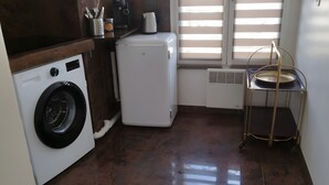 A compact kitchen with a washing machine, a small refrigerator, and a window with blinds.