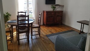 A living room with a wooden dining table, two chairs, a TV on the wall, a cabinet, and a sofa.