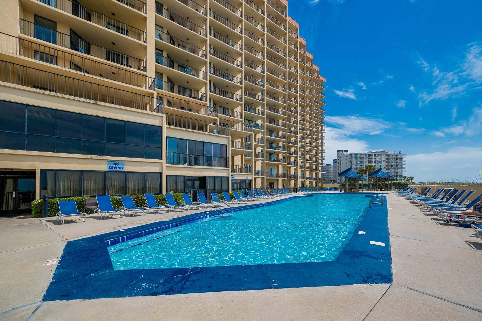 A hotel with a large swimming pool, multiple balconies, and a clear blue sky.