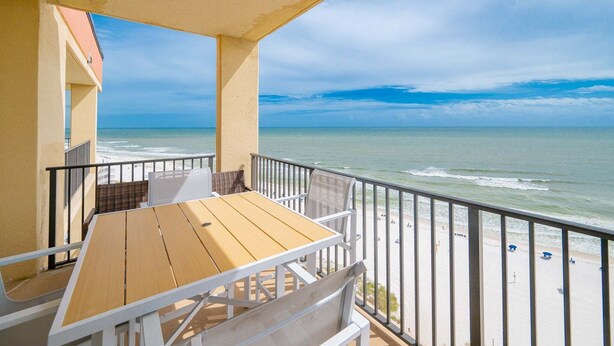 A balcony with a wooden table and chairs, overlooking a beach with the ocean and a clear sky. - Phoenix V 1516 2 Bedroom Condo (Orange Beach)