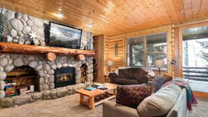 A log cabin interior with a stone fireplace, wooden furniture, and a large window. - Deer Valley Condo Near Base Hot Tub (Park City)