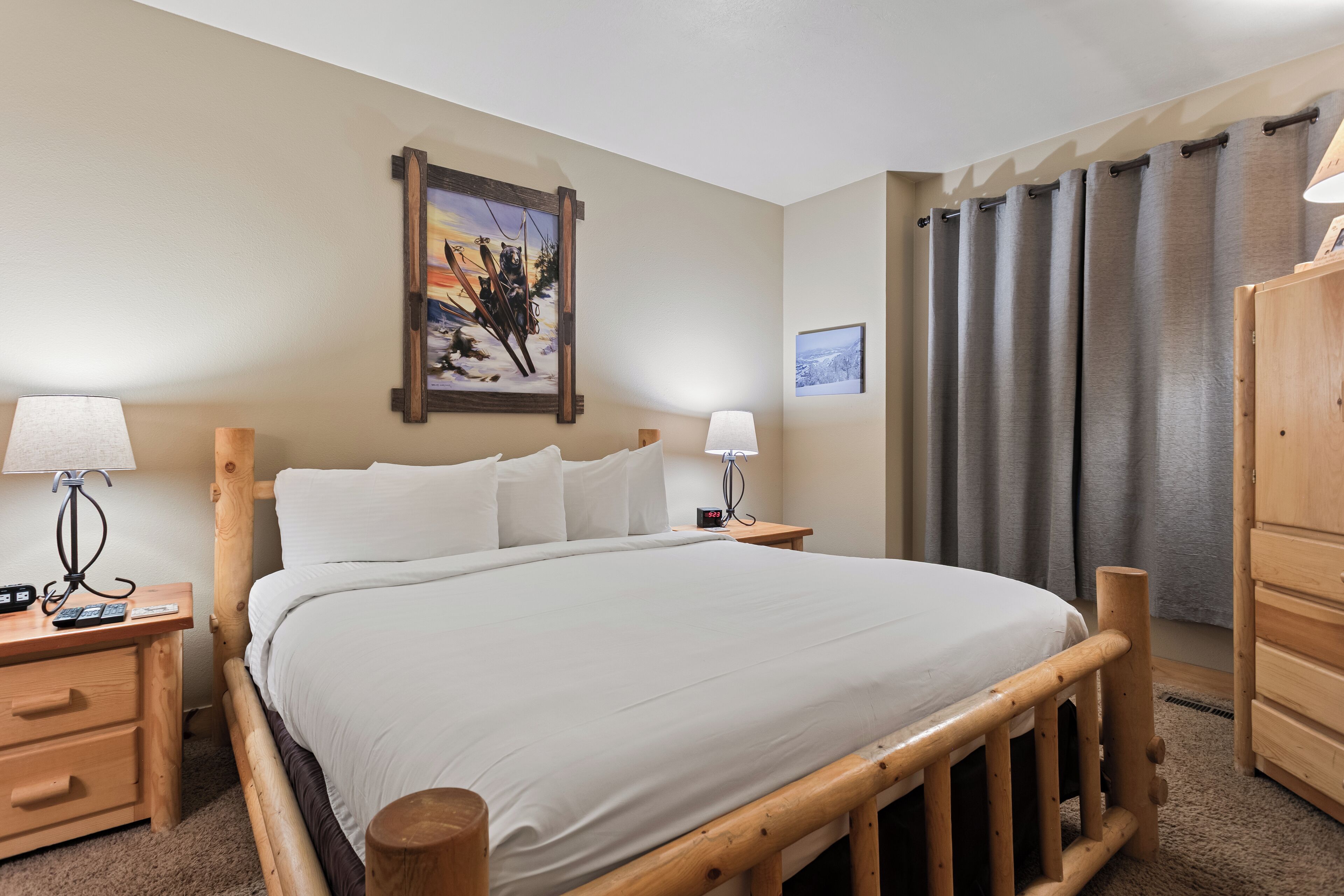 Een netjes opgemaakt bed met witte lakens, twee nachtkastjes met lampen, een houten hoofdbord, een houten garderobe en een schilderij aan de muur.