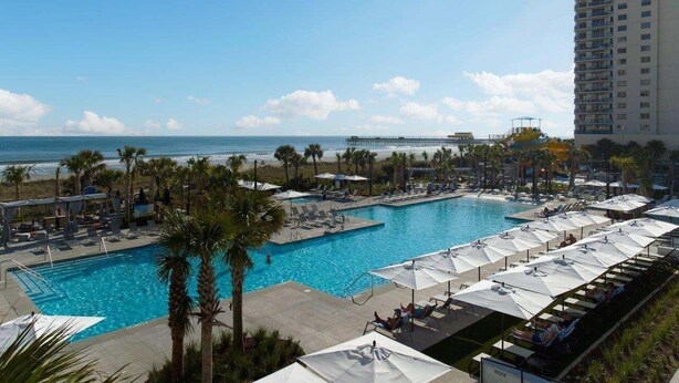 A resort pool with lounge chairs and umbrellas, a beach view, and a tall building in the background. - Windermere Coastal Retreat 7c 2 Bedroom Condo (Myrtle Beach)