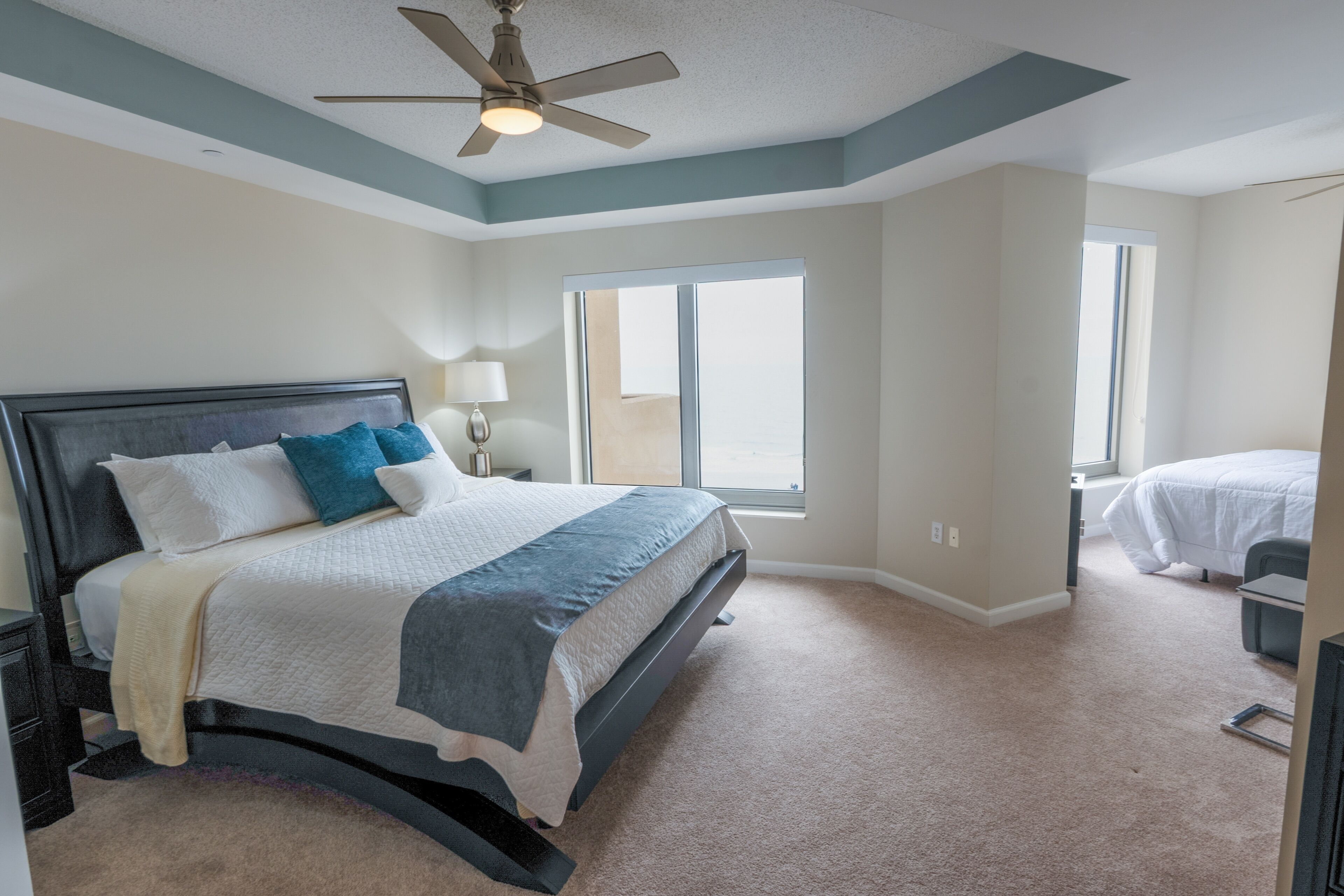 A bedroom with a bed, a nightstand, a lamp, a ceiling fan, and large windows.