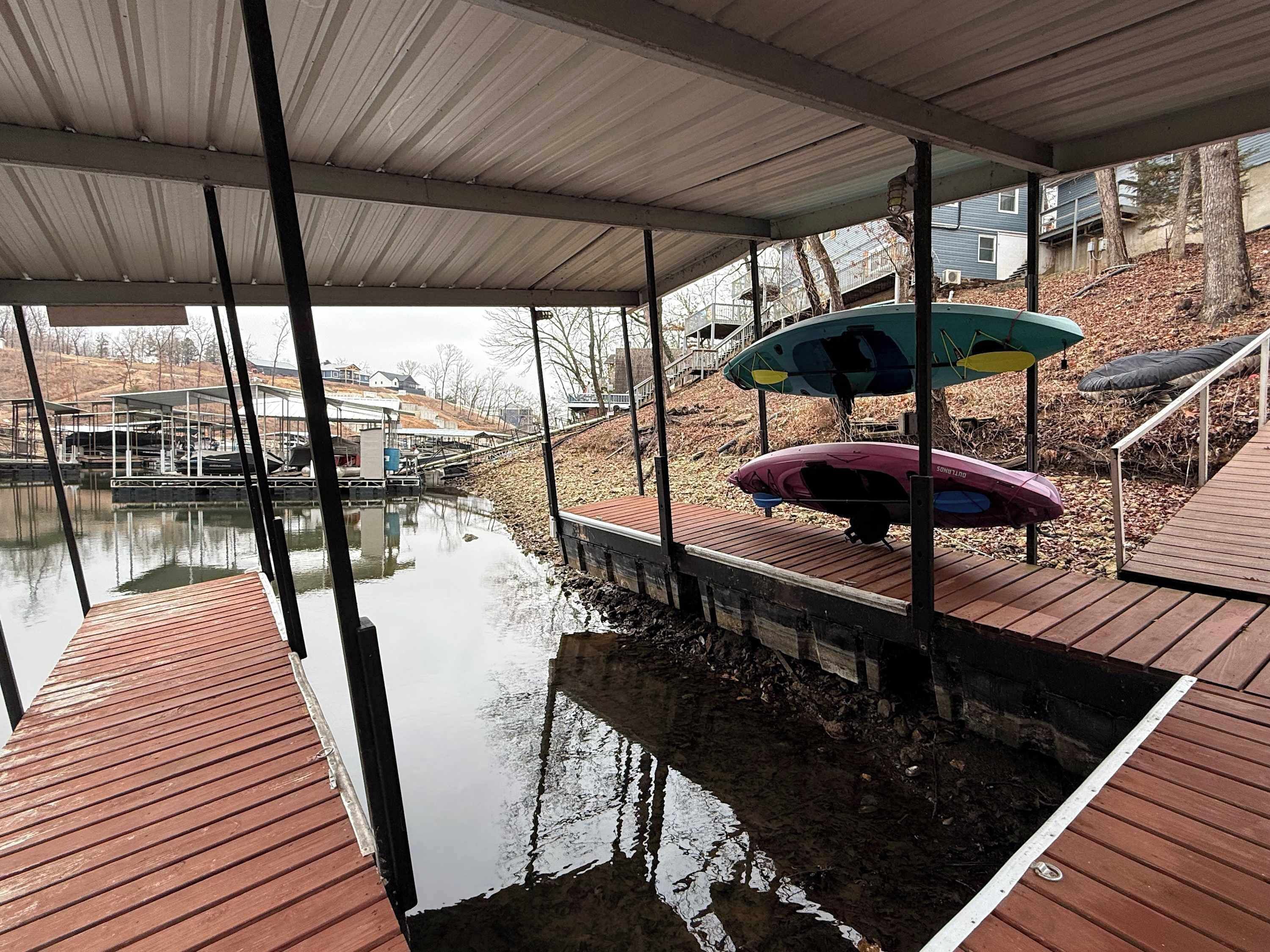 A covered dock area with a boat and a kayak, a calm body of water, and a view of houses in the background.