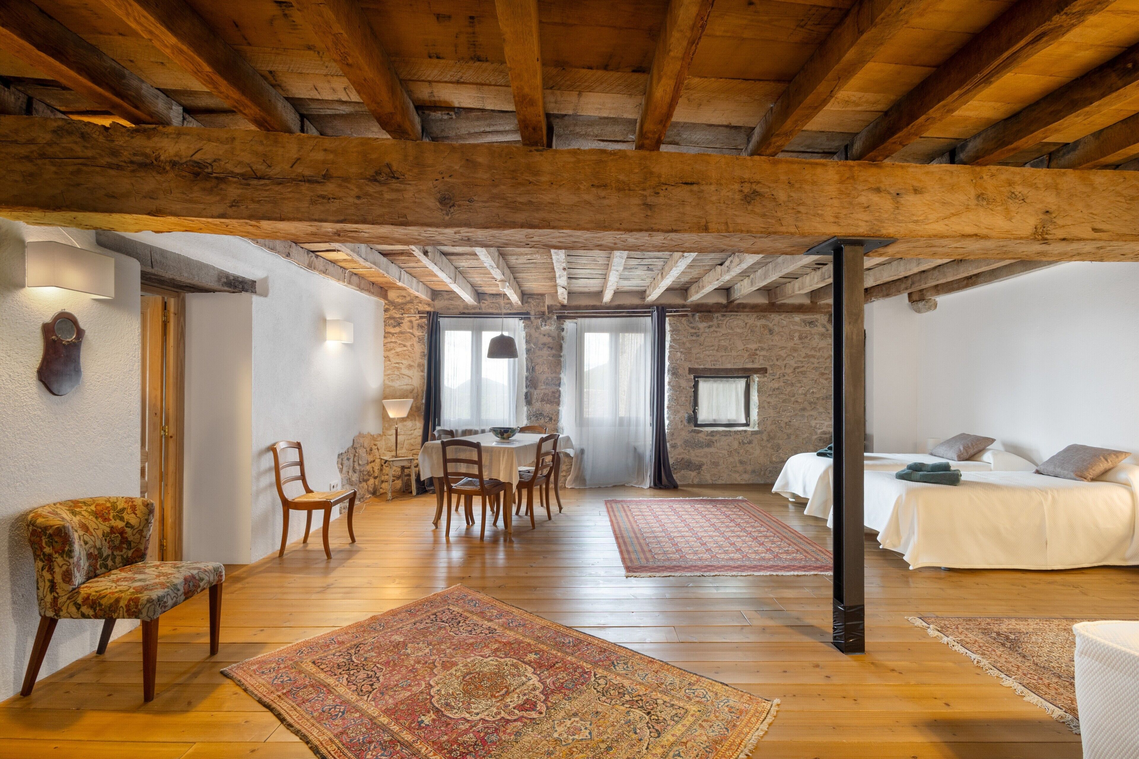 Un interior rústico con vigas de madera a la vista, una zona de comedor con sillas y una mesa, una cama con almohadas, y una alfombra estampada.