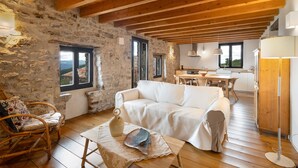Un interno rustico con pareti in pietra, travi in legno, un divano bianco, poltrone in vimini, un tavolo da pranzo e un angolo cottura.