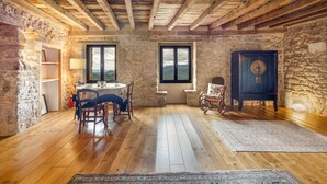 Una stanza rustica con pareti in pietra, travi in legno, un tavolo rotondo con sedie, una sveglia e vista sull'esterno.