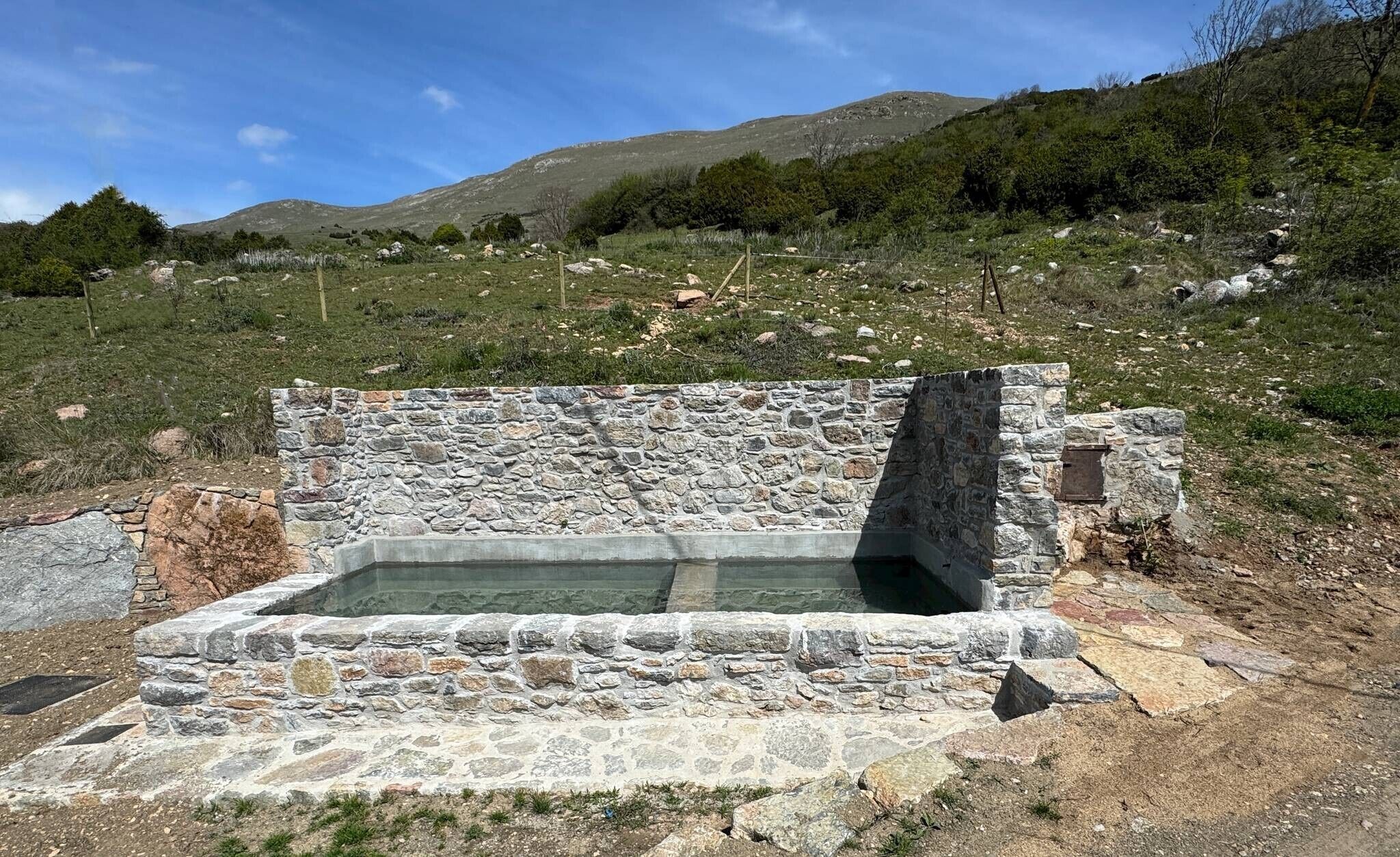 Una struttura in pietra con una piscina rettangolare, circondata da un muro in pietra, immersa in un paesaggio collinare.