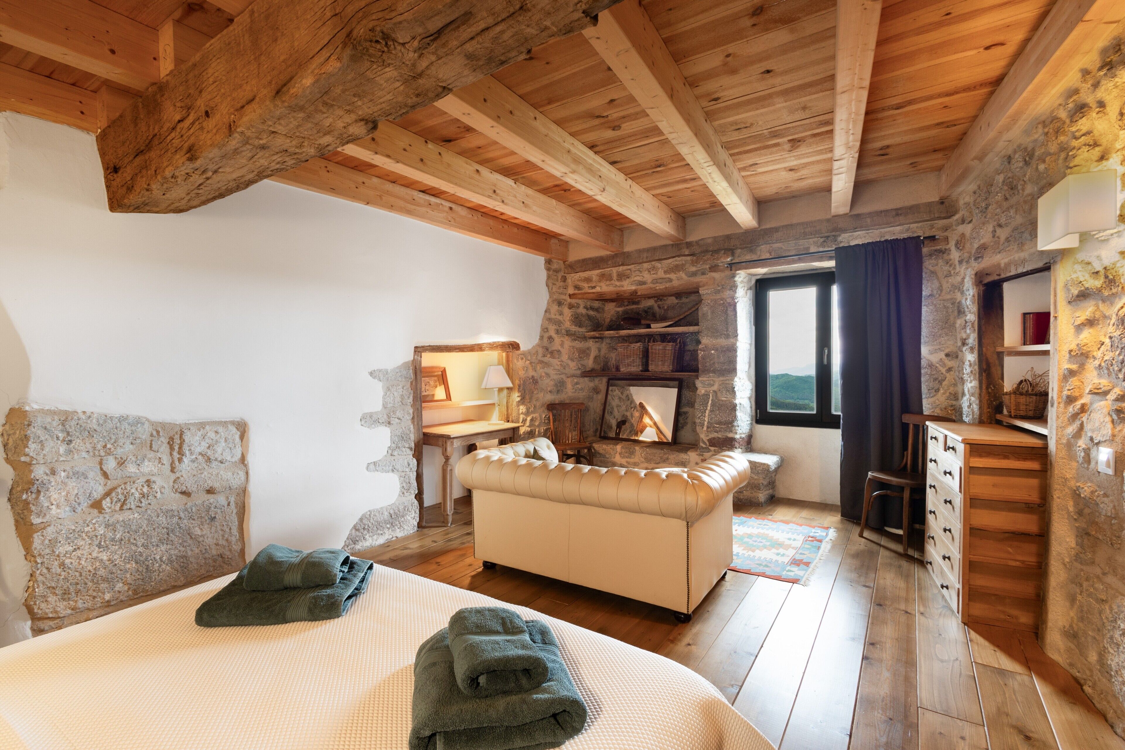 A bedroom with a bed, a sofa, a dresser, a stone wall, a fireplace, and a window.