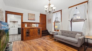 Un soggiorno con pavimento in legno, un divano grigio, un lampadario e un angolo cucina con mobili bianchi.