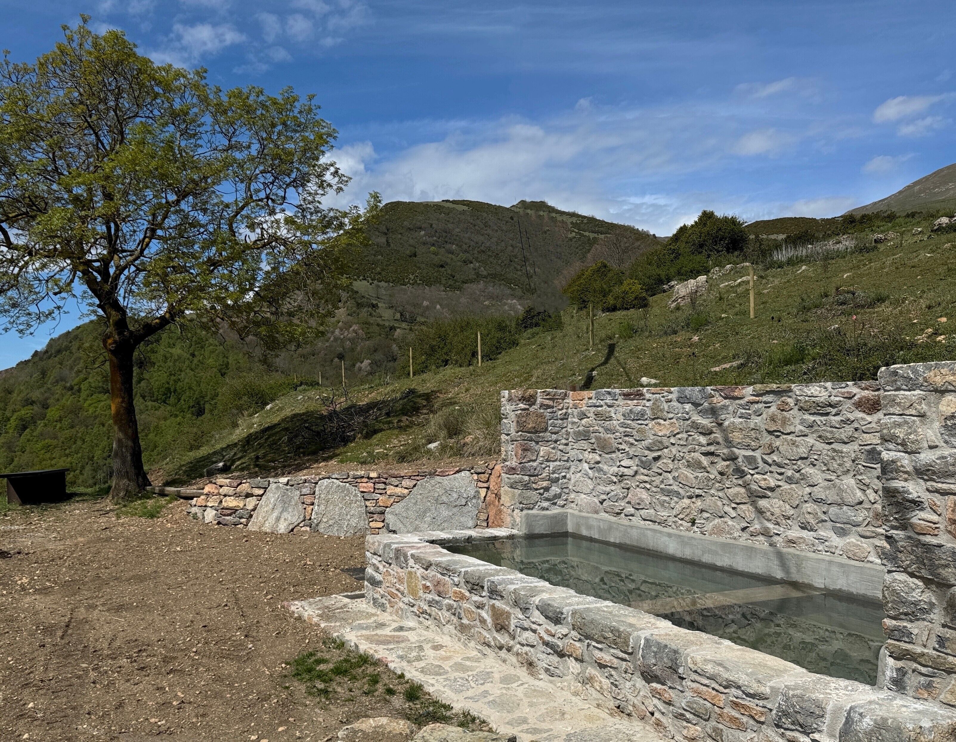A stone structure with a pool, surrounded by a stone wall, trees, and a hilly landscape.
